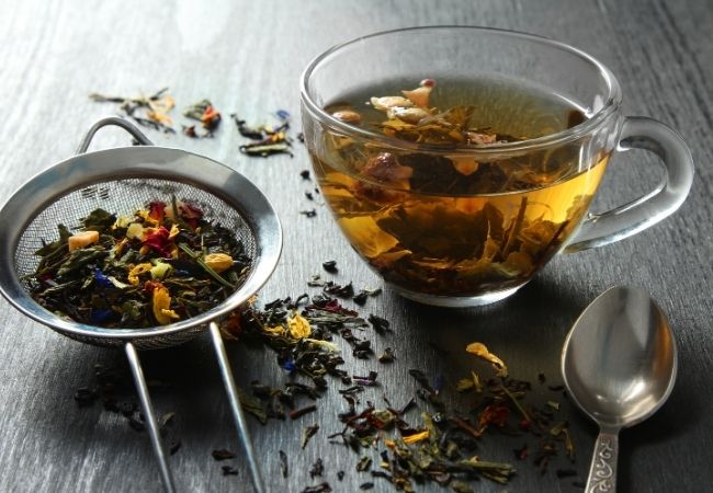 Herbs and dried flowers in a tea strainer and a cup of tea in a clear glass mug