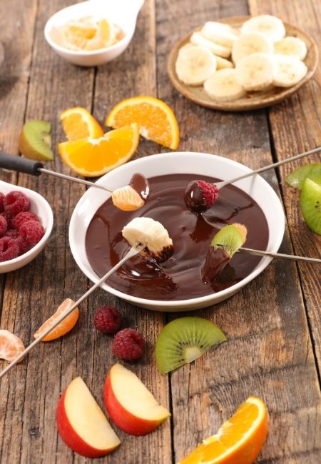 Chocolate fondue with fondue forks holding kiwi, apple wedges, orange wedges, bananas, and strawberries 