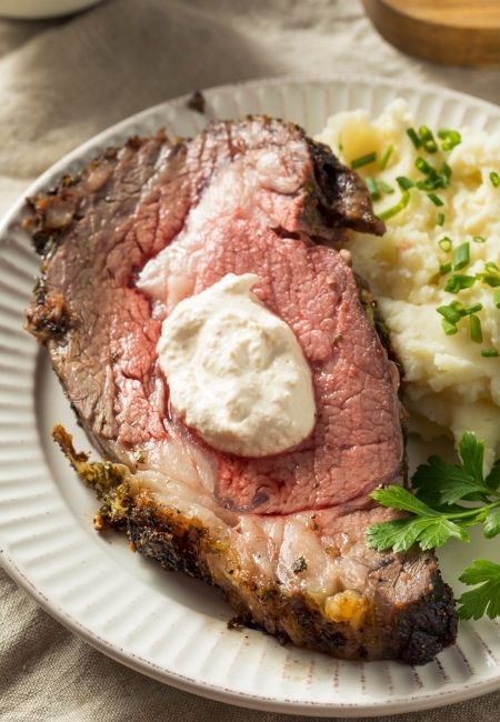Slice of Prime Rib on white plate with creamy horseradish sauce on top with side of mashed potatoes topped with green herbs 